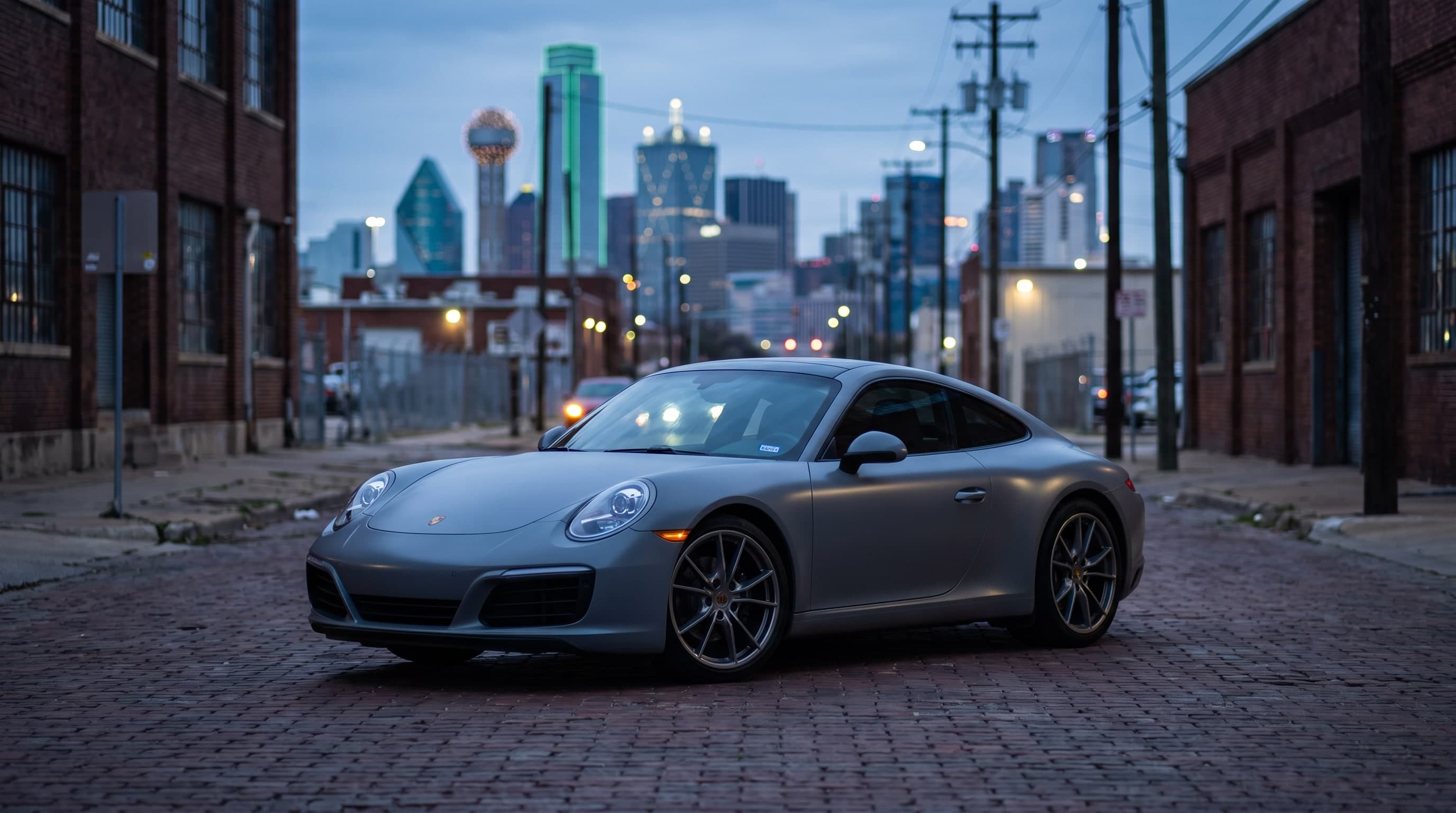 Satin grey wrapped Porsche 911 parked on a Design District street with the Dallas skyline behind it at dusk