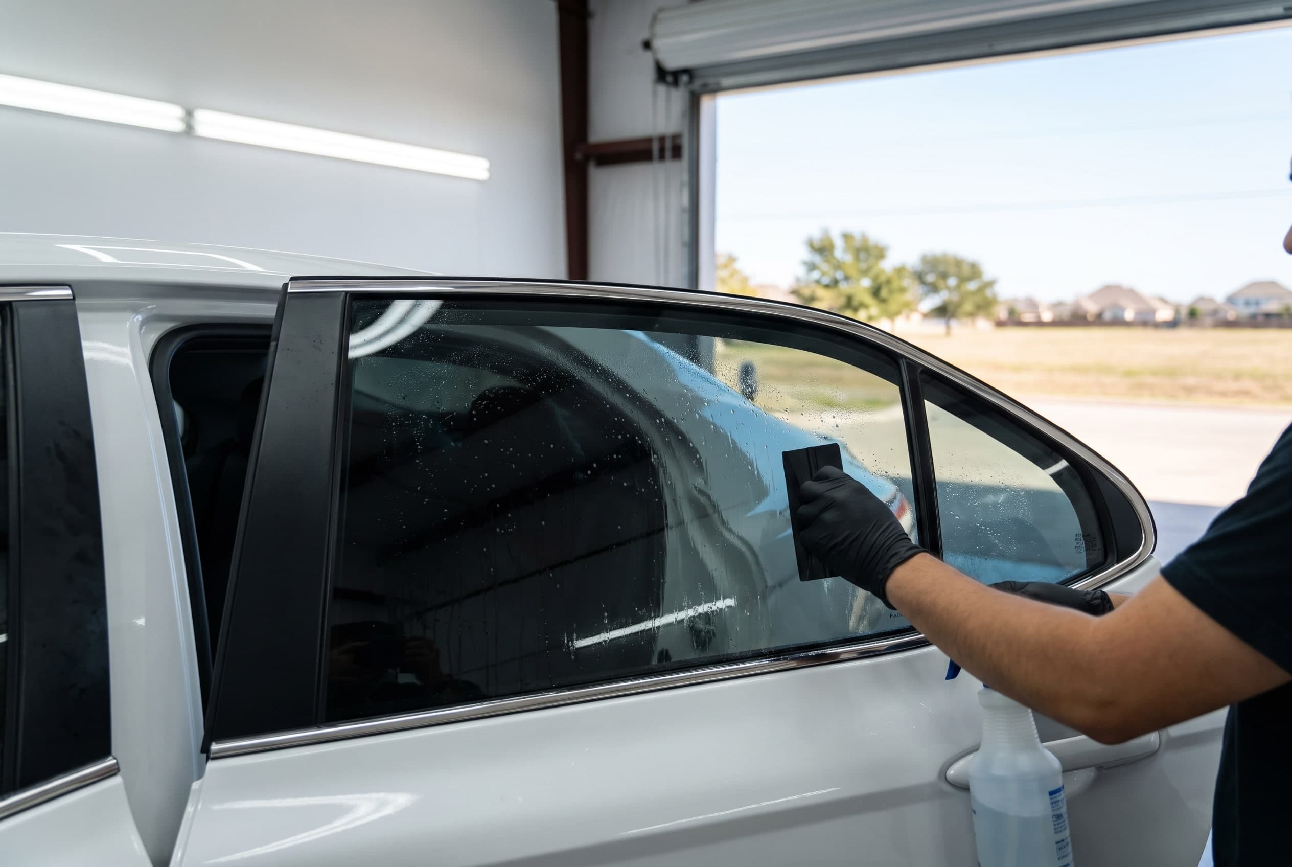 Technician squeegeeing ceramic window film onto the rear door glass of a white Toyota Camry inside a tint bay in Irving, TX